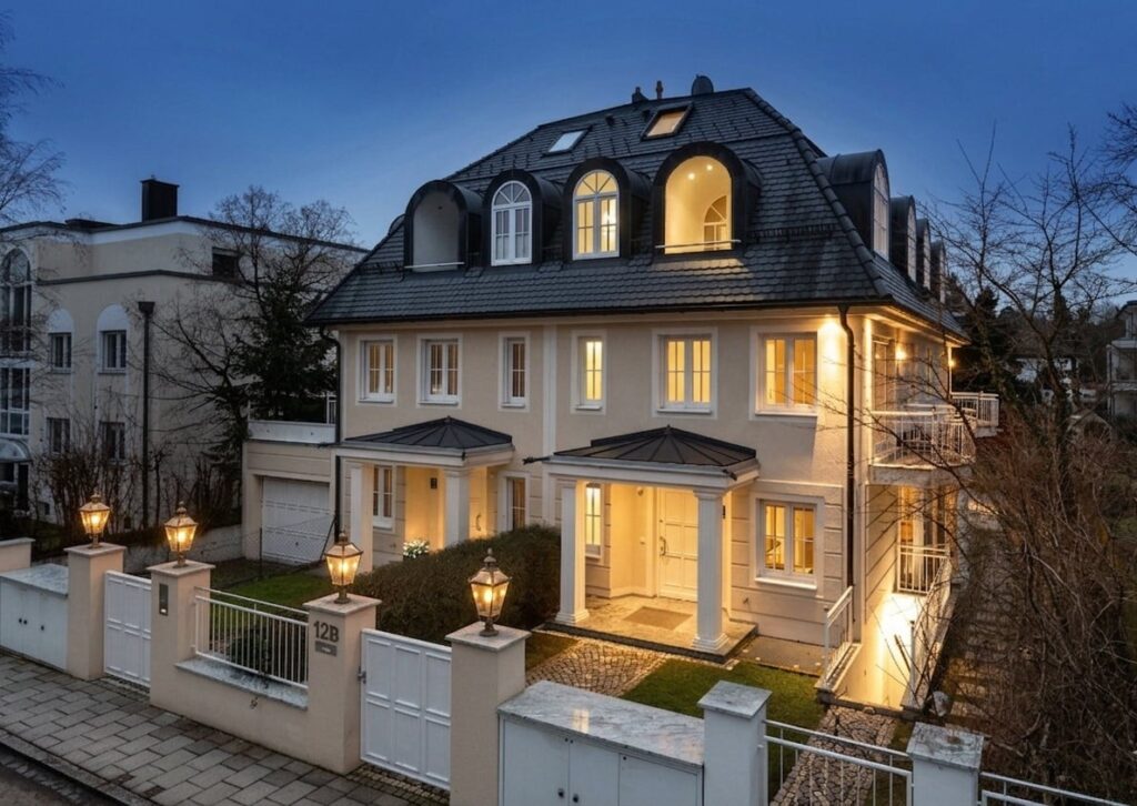 Großes, mehrstöckiges Wohnhaus mit beleuchteten Fenstern, Mansardendach und weißem Zaun in einem Wohnviertel bei Dämmerung