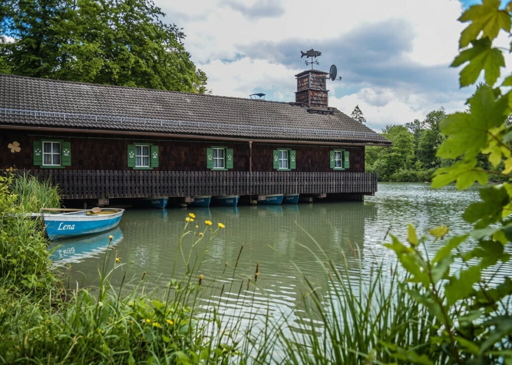 Holzhaus mit grün lackierten Fensterläden auf Pfählen über einem See mit einem blauen Ruderboot namens 'Lena' am Ufer