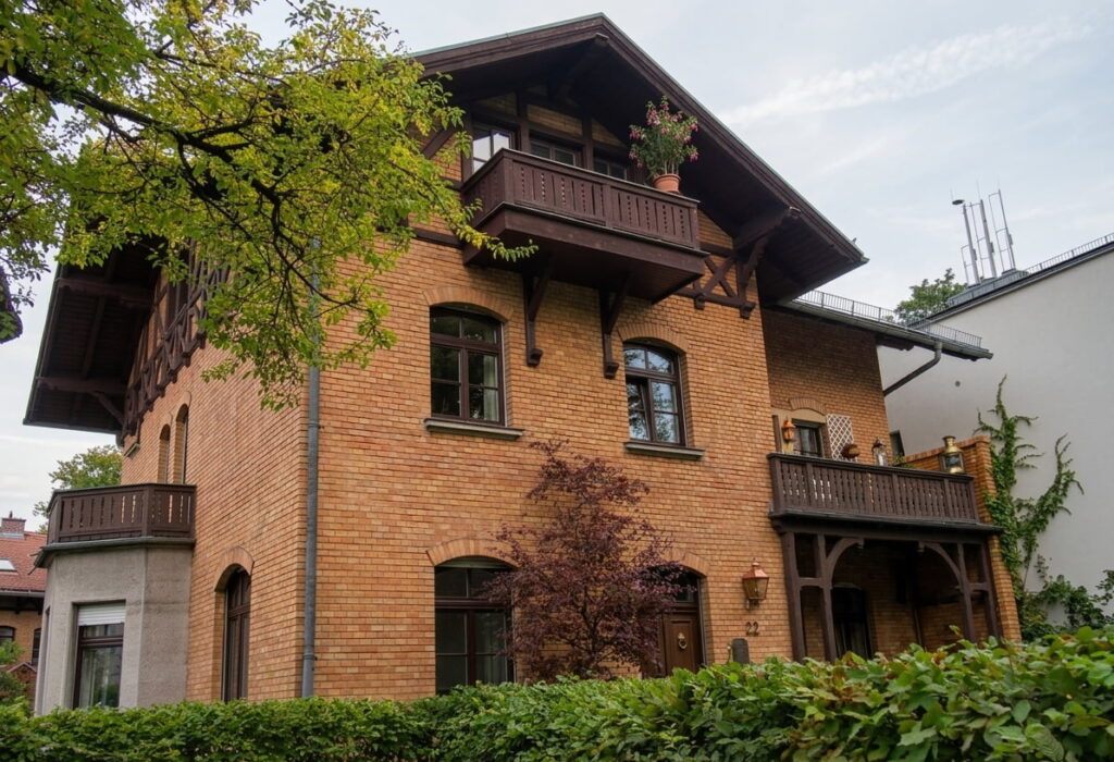 Mehrstöckiges Backsteinhaus mit zwei hölzernen Balkonen, umgeben von Bäumen und Sträuchern